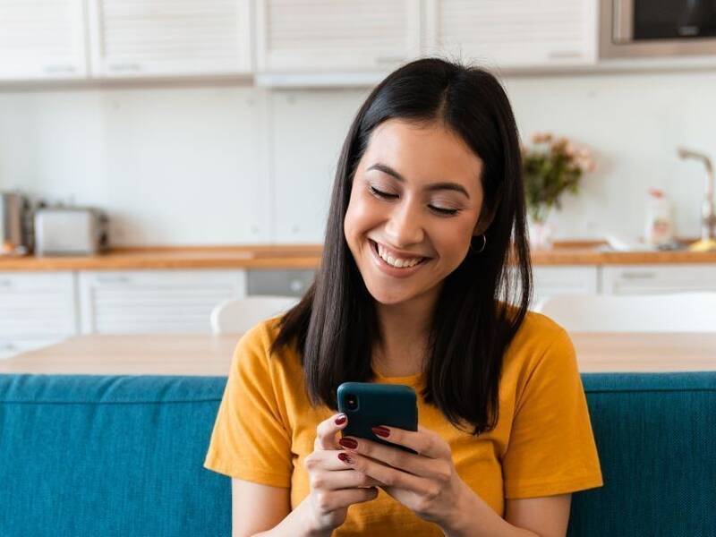 Hier ist eine Frau zu sehen, die ein Handy in ihren beiden Händen hat, auf einem Sofa sitzt und etwas am Smartphone erledigt. Im Hintergrund ist einen Küche. 