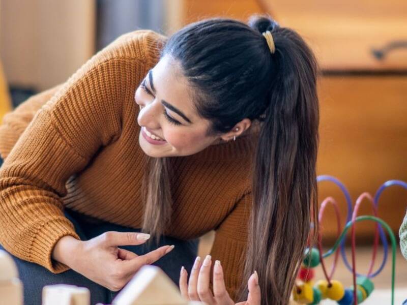 Hier ist einen junge Frau zu sehen, die ein Kind anlacht und dabei mit ihm spielt. Im Hintergrund sin d auch noch Kinder.