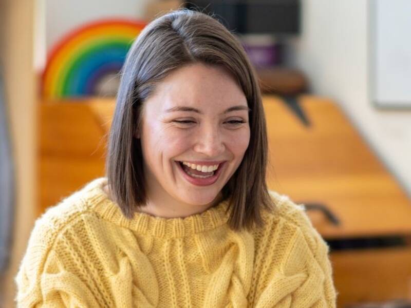 Hier ist eine lachende Frau in einem gelben Pulli zu sehen.