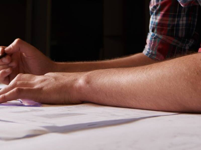 Hier sind zwei Unterarme von einem Mann zu sehen. Sie liegen auf einem Plan und die Hände halten Bleistift und Dreieck.