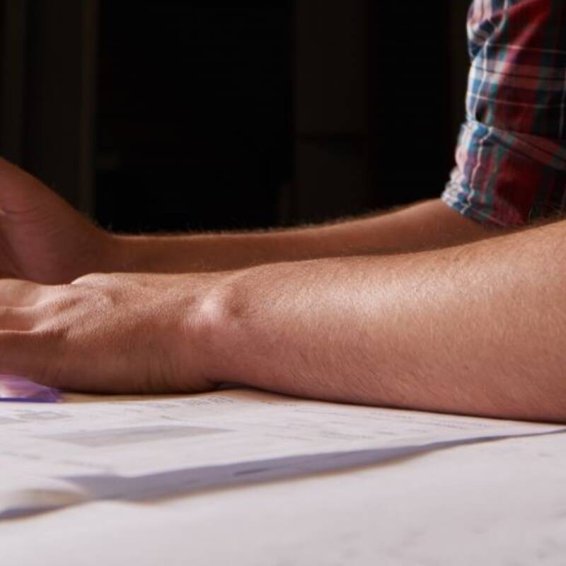 Hier sind zwei Unterarme von einem Mann zu sehen. Sie liegen auf einem Plan und die Hände halten Bleistift und Dreieck.