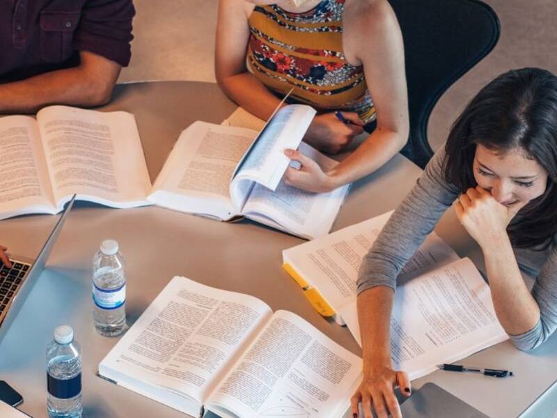 Hier sind junge Leute an einem Tisch zu sehen. Auf dem Tisch sind Bücher und ein Laptop. Sie lernen wahrscheinlich.