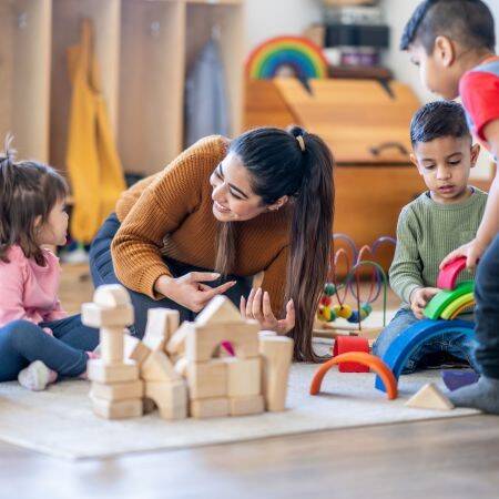 Hier ist eine frau zu sehen, die mit einem Kind spielt. Im Vordergrund stehen Holzbauklötze und daneben sind zwei weiter Kinder, die spielen. 