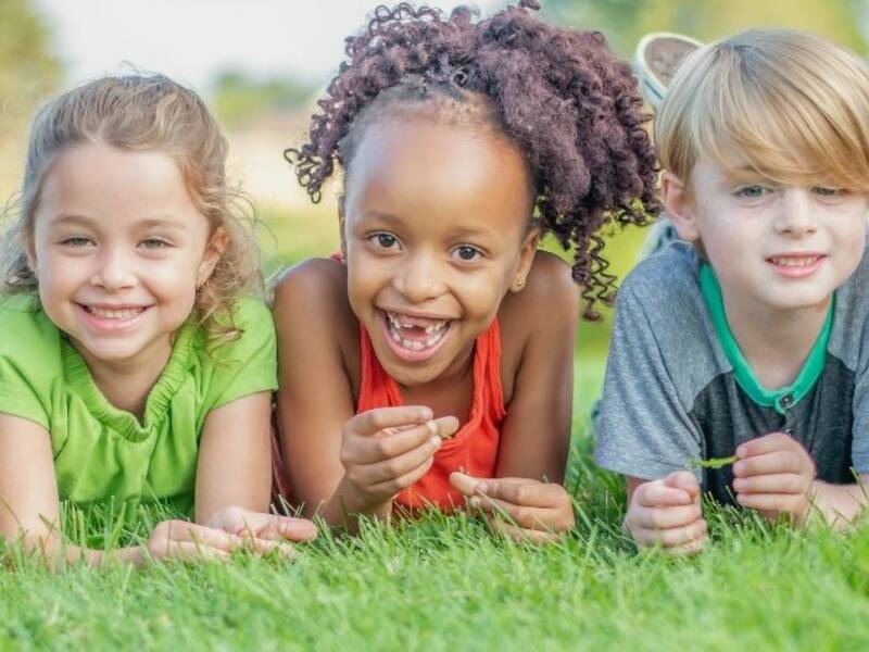 Hier sind Kinder zu sehen, die auf dem Bauch im Gras liegen und den betrachter anschauen.