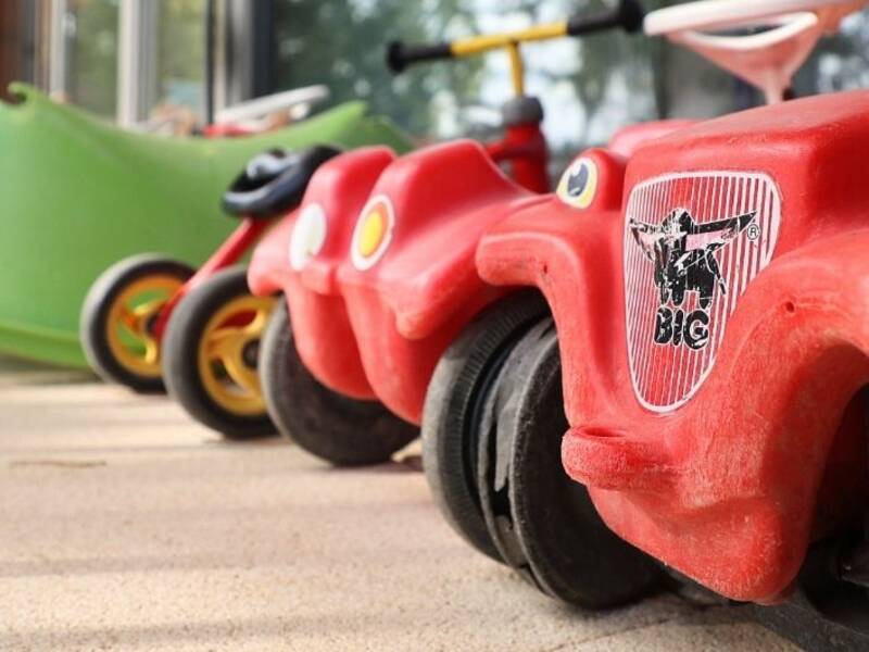 Hier isein rotes Bobby-Car im Vordergrund rechts zu sehen. Es steht in einer Reihe mit anderen Bobby-Cars in rot und grün. 