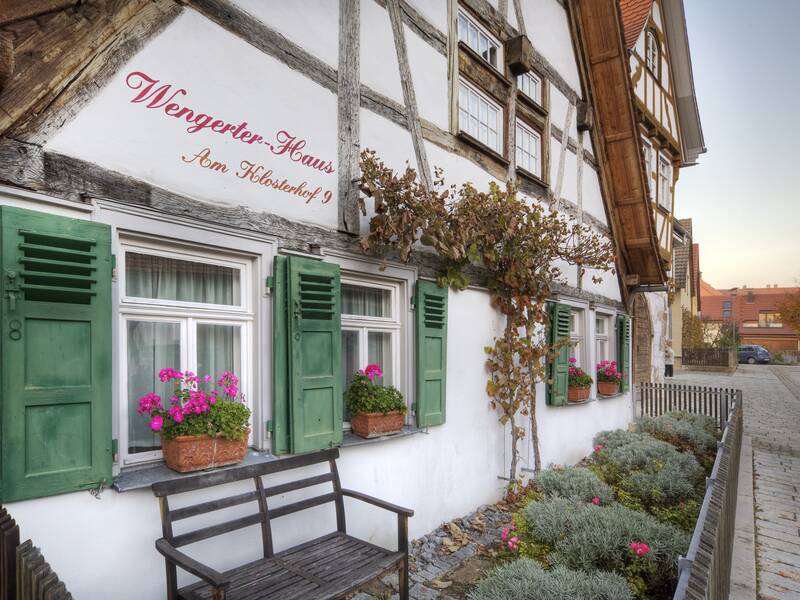 Ein Fachwerkhaus mit der Aufschrift "Wengerter-Haus Am Klosterhof 9" hat grüne Fensterläden, blühende Blumenkästen und eine Bank davor.