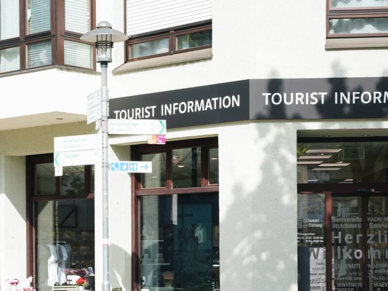 Das Haus, in dem die Touristeninformation ist, ist zu sehen. Vor der MMT Metzingen steht ein rosa geschmücktes Fahrrad sowie ein Tisch und zwei einfache Stühle in grün. Die Schaufenster des Verkaufsraums ist zu sehen. Es ist ein sonniger Tag.