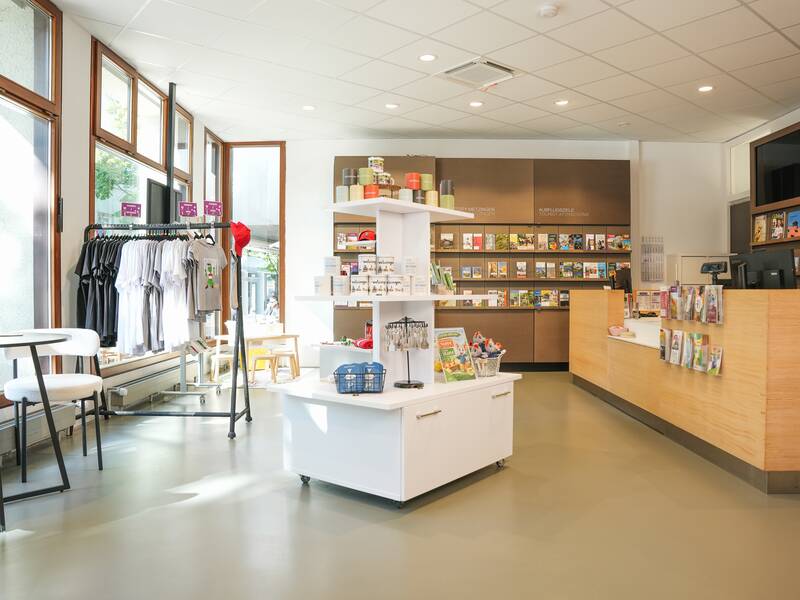 Zu sehen ist der Verkaufsraum der MMT Metzingen mit Theke, Bücherregal, Klamottenstange und Souvenirtisch in der Mitte. Das Angebot der MMT ist zu sehen.