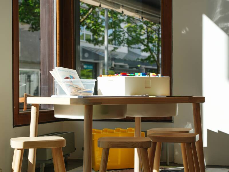 Im Verkaufsraum der MMT Metzingen ist der Kinderspielbereich zu sehen. Es gibt einen kleinen Holztisch mit Spielzeug und zwei Holzhocker. Der Spielbereich ist direkt am Fenster, durch das die Sonne scheint.