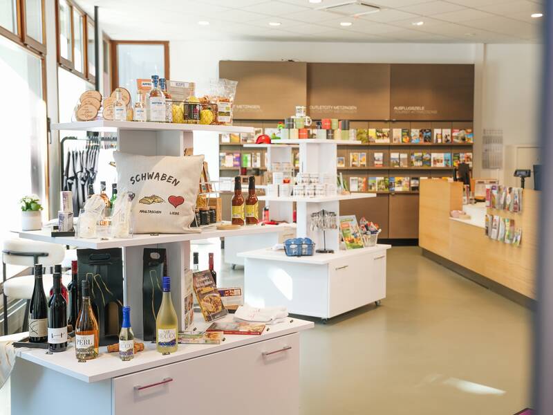 Zu sehen ist der Verkaufsraum der MMT Metzingen mit Theke, Bücherregal, Klamottenstange und Souvenirtisch in der Mitte. Das Angebot der MMT ist zu sehen.