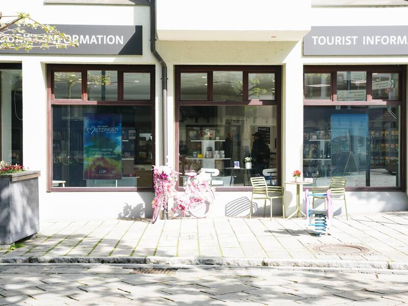 Vor der MMT Metzingen steht ein rosa geschmücktes Fahrrad sowie ein Tisch und zwei einfache Stühle in grün. Die Schaufenster des Verkaufsraums ist zu sehen. Es ist ein sonniger Tag.