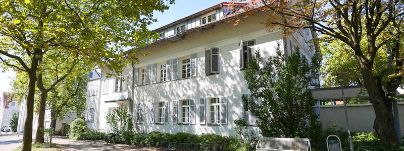 Musikschule. Ein zweistöckiges, weißes Gebäude mit Sprossenfenstern steht von Bäumen umgeben an einem sonnigen Tag.