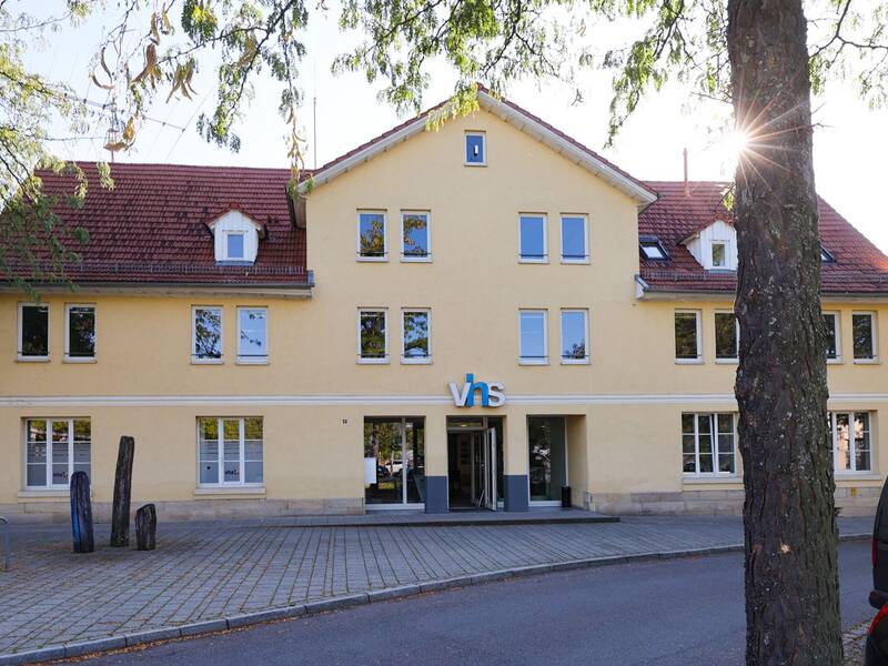 Volkshochschule. Ein gelbes Gebäude mit roten Ziegeldach und dem Schriftzug "vhs" über dem Eingang steht hinter einem Baum, während die Sonne durch die Äste scheint.