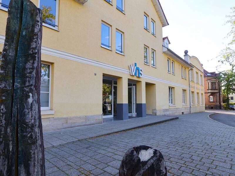 Volkshochschule. Ein gelbes Gebäude mit der Aufschrift "vhs" steht an einem Gehweg, umgeben von Bäumen und einem Holzobjekt im Vordergrund.