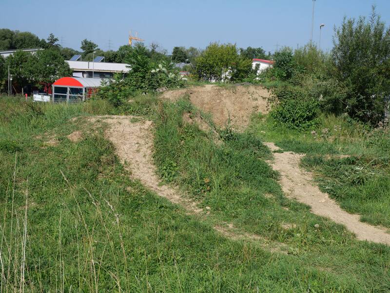 Eine von Gras und Büschen bewachsene Erdpiste verläuft durch eine grüne Hügellandschaft, im Hintergrund sind moderne Gebäude zu sehen.