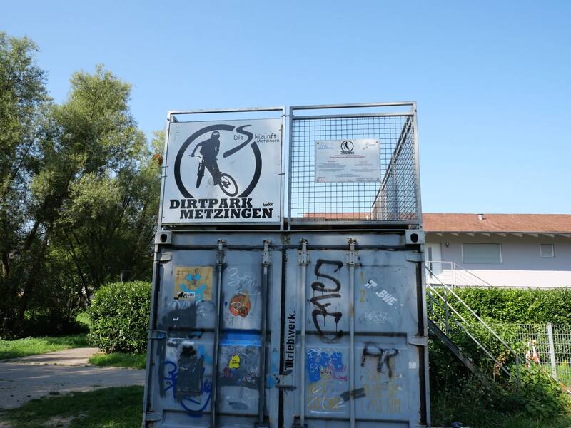 Vor einem klaren blauen Himmel steht ein grauer Container mit Grafitti, auf dem ein Schild mit der Aufschrift "Dirtpark Metzingen" und der Silhouette eines Radfahrers angebracht ist.