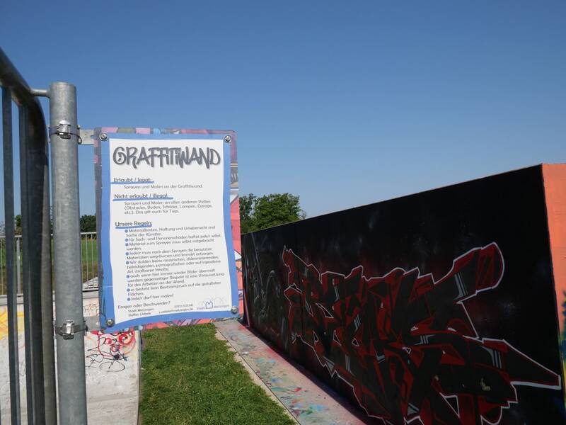 Ein Schild erklärt die Regeln für die legale Graffitiwand, darunter erlaubte und verbotene Aktivitäten. Daneben beginnt die bemalte schwarze Wand mit rotem Graffiti, im Hintergrund Rasen und blauer Himmel.