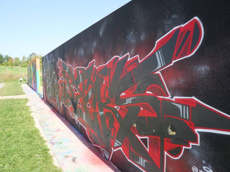 Eine schwarze Wand mit rotem und grauem Graffiti-Schriftzug, umgeben von einer gesprenkelten Sternenhimmel-Optik. Der Boden davor ist mit bunten Farbflecken übersät, im Hintergrund ist Wiese und blauer Himmel zu sehen.