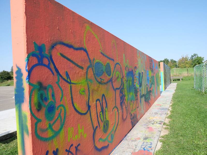 Die lange, orangefarbene Graffitiwand zieht sich in die Tiefe. Darauf sind bunte Comicfiguren und Kritzeleien in Blau, Grün und Gelb zu sehen. Rechts daneben verläuft ein Zaun, im Hintergrund grüne Wiese.