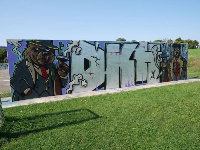 Ein aufwendig gestaltetes Graffiti zeigt zwei Bären in Anzügen mit Maschinenpistolen, flankierend zu einem großen silbernen Schriftzug. Im Hintergrund blauer Himmel und grüne Wiese.