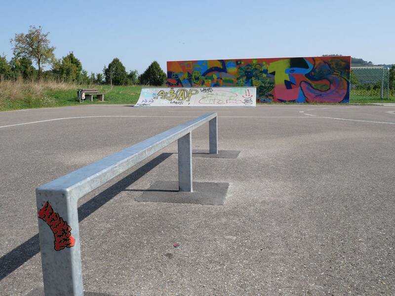 Eine lange, niedrige Metallstange auf Asphalt zum Grinden, im Hintergrund eine bunte Graffiti-Wand.