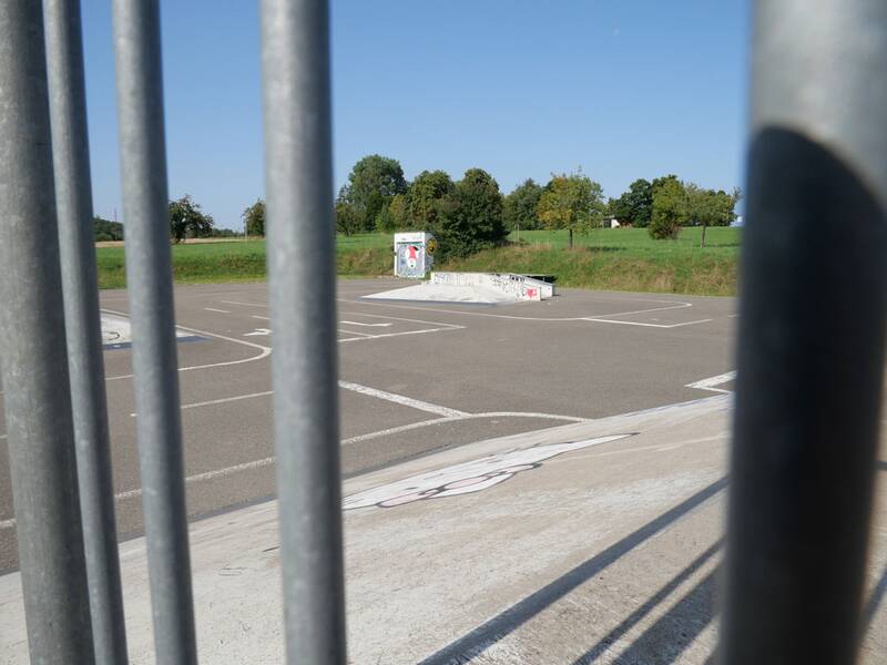 Blick durch Metallgitter auf die Skateanlage, im Hintergrund steht eine mit Graffiti bemalte Quarterpipe.