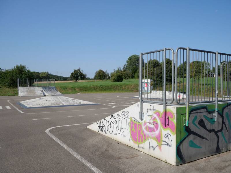 Skateanlage mit mehreren Rampen und Hindernissen auf einer asphaltierten Fläche vor grüner Wiese.