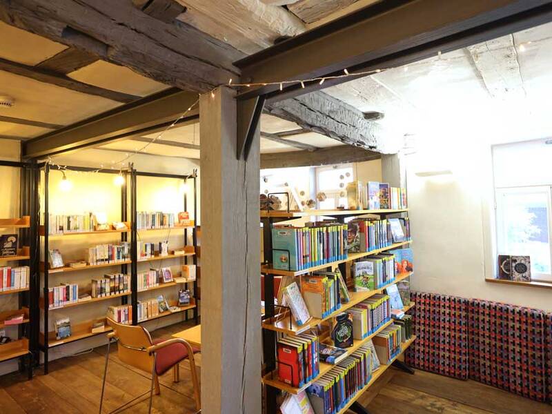 Ein gemütlicher Raum mit Holzbalken-Decke zeigt Regale voller Bücher, eine gepolsterte Sitzbank unter einem Fenster und einen einzelnen, roten Stuhl.