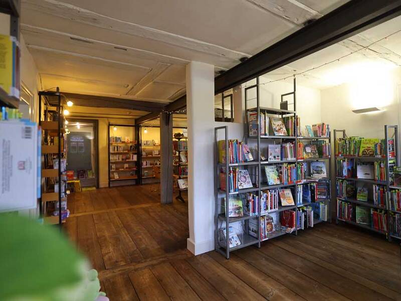 Ein Raum mit hölzernem Boden zeigt mehrere Regale voller Bücher, davon sind einige Kinderbücher, in einem hell beleuchteten, gemütlichen Buchladen.