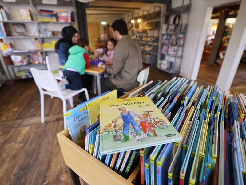 Es sind mehrere Bücher zu sehen, während im Hintergrund eine Familie um einen Tisch zusammensitzt.