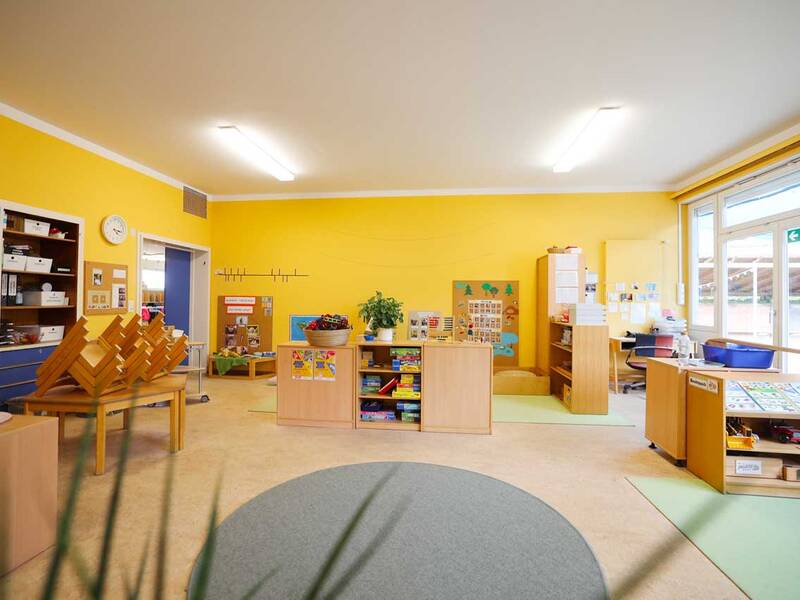 Ein heller, geordneter Kindergartenraum mit gelben Wänden, Tischen, Regalen voller Bücher und Spielzeug sowie Fenstern, die viel Licht hereinlassen.