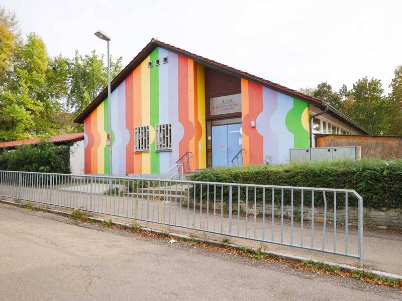 Ein Gebäude mit einem farbenfrohen, vertikal gestreiften Design in bunten Farben steht hinter einem Metallzaun, umgeben von grünen Bäumen.