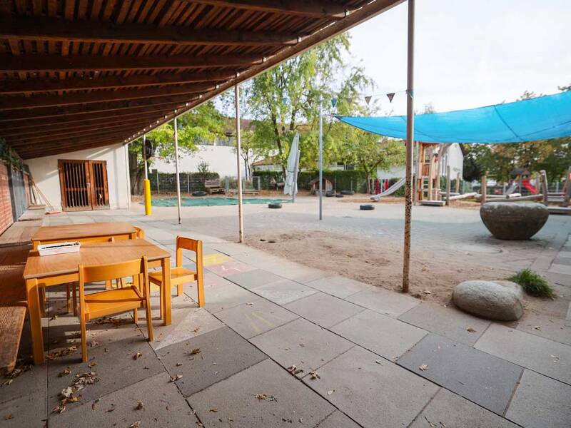 Der Außenbereich eines Kindergartens enthält Holztische und -stühle unter einem Vordach sowie Spielgeräte im Hintergrund.