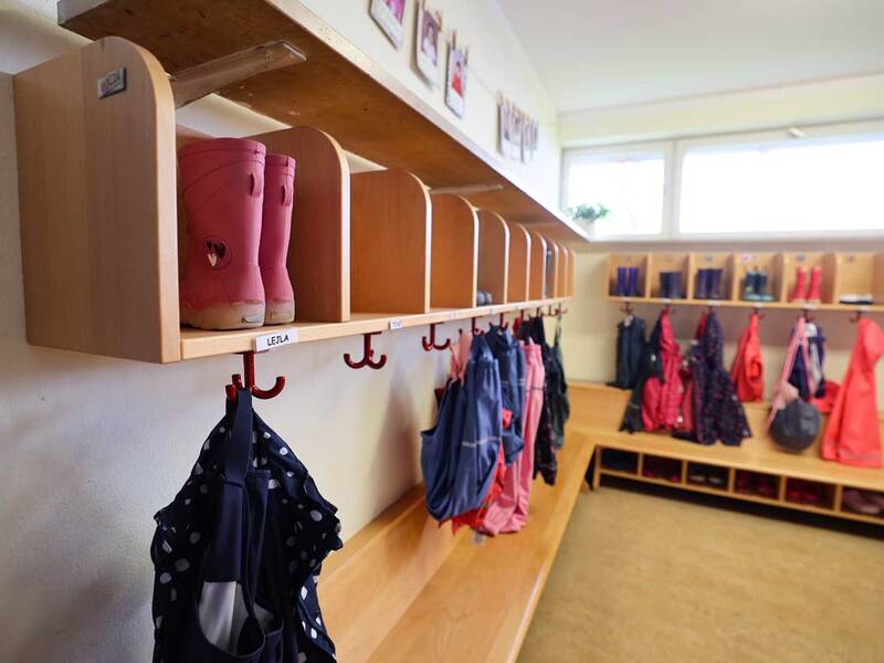 Ein Kindergarten-Garderobenbereich mit Haken und Fächern, an denen bunte Kinderregenjacken und Gummistiefel hängen und stehen.