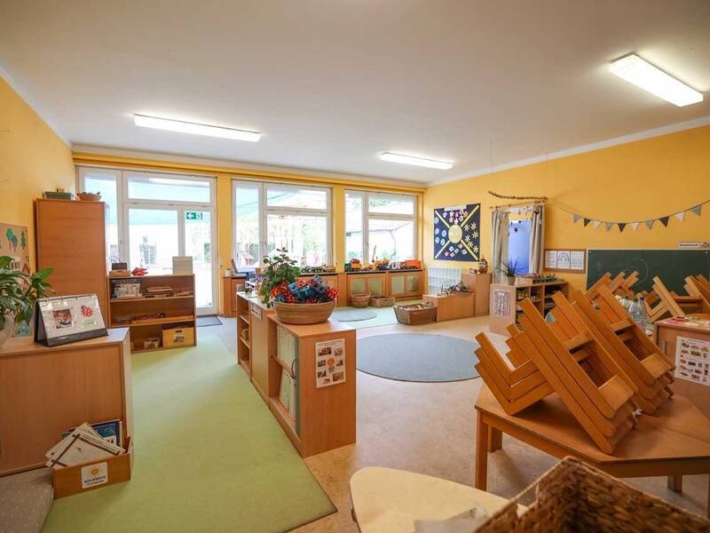 Ein gut beleuchteter Kindergartenraum mit gelben Wänden, Holzmöbeln, Grünpflanzen und Spielzeug, wobei einige Stühle auf den Tischen gestapelt sind.