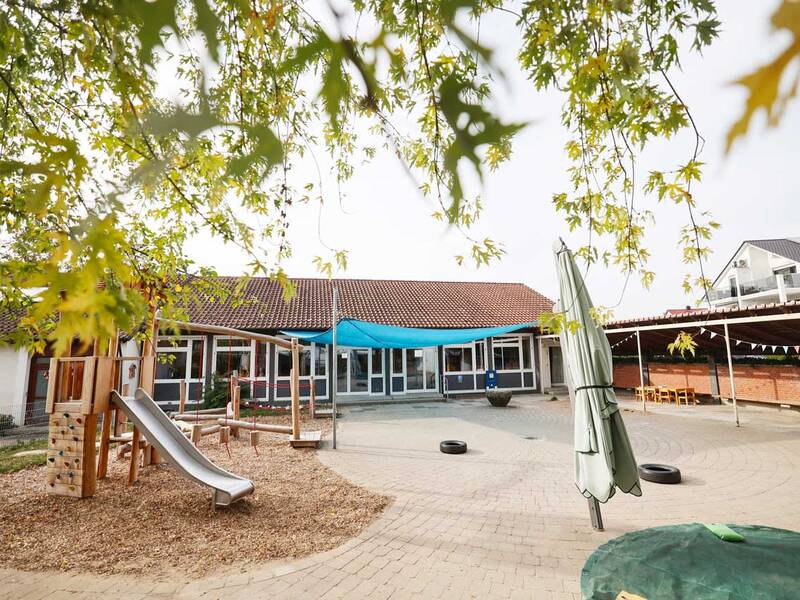 Ein Spielplatz mit Holzklettergerüst und Rutsche befindet sich vor einem Gebäude mit rotem Ziegeldach, umgeben von grünem Laub.