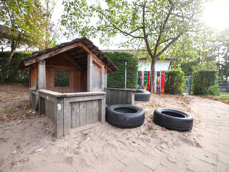 Ein kleiner hölzerner Sandkasten steht auf einem Spielplatz unter einem Baum, umgeben von alten Reifen und herbstlichem Laub.