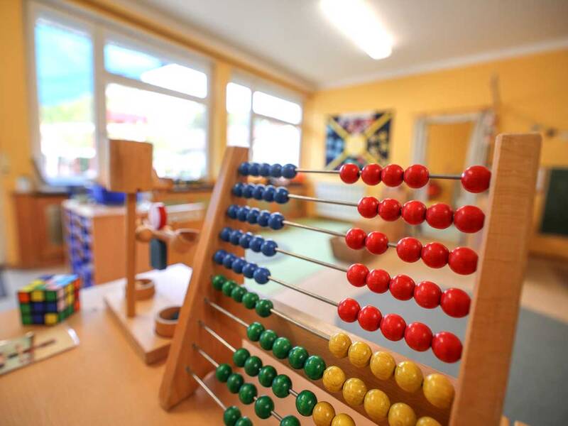 Ein bunter Abakus mit roten, blauen, grünen und gelben Kugeln steht auf einem Tisch in einem hellen Raum mit gelben Wänden, umgeben von weiteren Spielzeugen.