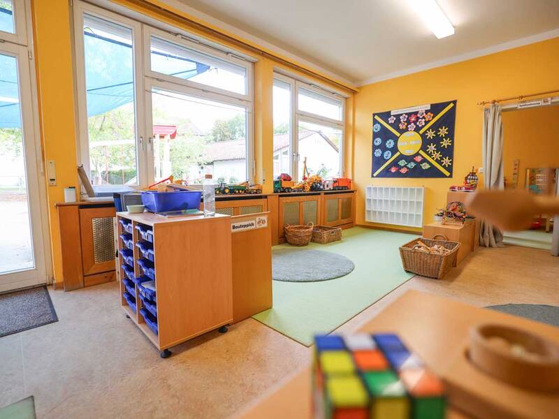 Ein Kindergartenraum mit gelben Wänden, großen Fenstern, Spielzeugregalen und einem bunten Wandplakat mit verschiedenen Formen.