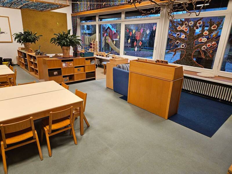 Ein heller Raum mit großen Fenstern zeigt ein Schulzimmer oder Kindergarten mit Tischen, Stühlen, Regalen und dekorativen Elementen an den Fenstern.