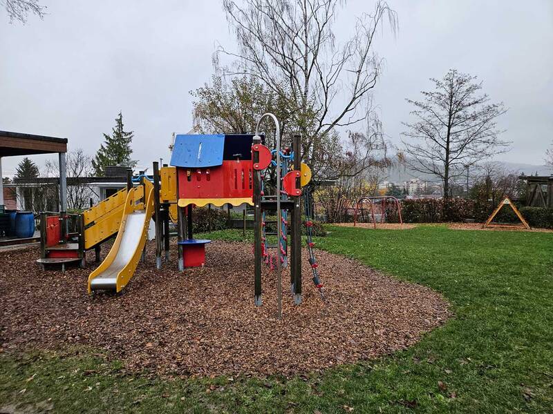 Ein Spielplatz mit bunten Klettergerüsten, einer Rutsche und Schaukeln steht auf einer Fläche mit Rindenmulch und Rasen, umgeben von Bäumen.