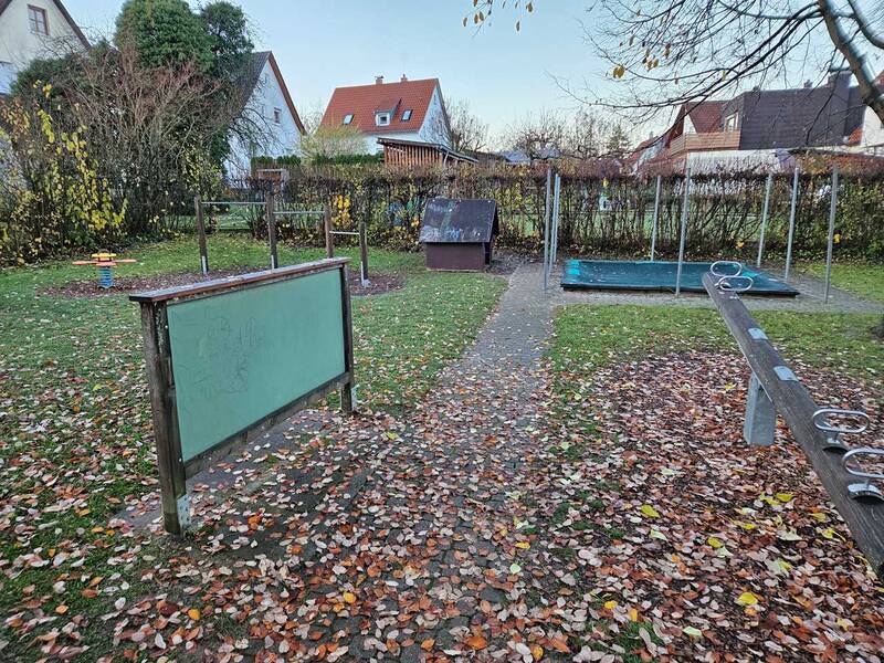Ein herbstlich mit Laub bedeckter Spielplatz mit Außentafel, Balancierbalken, Federtierwippe und Klettergerüst umgeben von Büschen und Häusern.