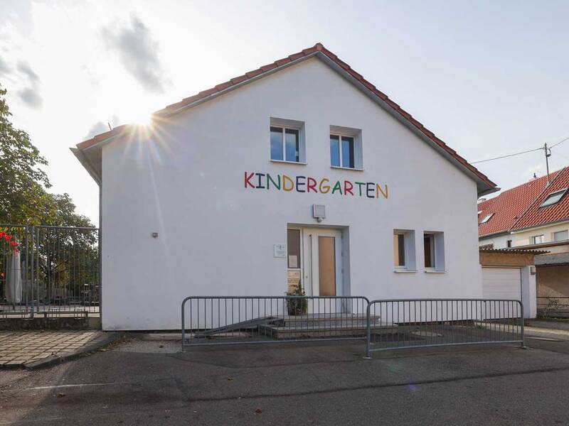 Ein weißes Gebäude mit der bunten Aufschrift "KINDERGARTEN" vorne und einem Gitterzaun im Vordergrund steht unter einem sonnigen Himmel.