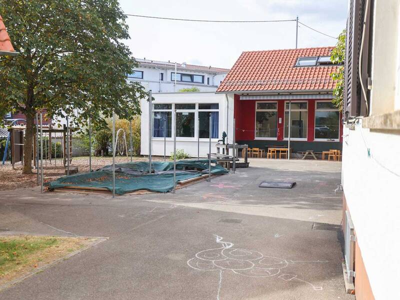 Der Außenbereich eines Gebäudes mit einem Spielplatz, Klettergerüsten, einem Baum und einem rot-weißen Haus mit angrenzenden Sitzgelegenheiten ist zu sehen.