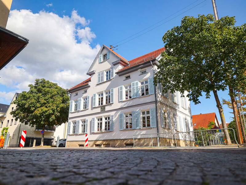 Ein dreistöckiges Gebäude mit weißen Wänden, hellblauen Fensterläden und rotem Dach. Es wirkt gepflegt und freundlich, steht an einer gepflasterten Straße und ist von Bäumen umgeben.