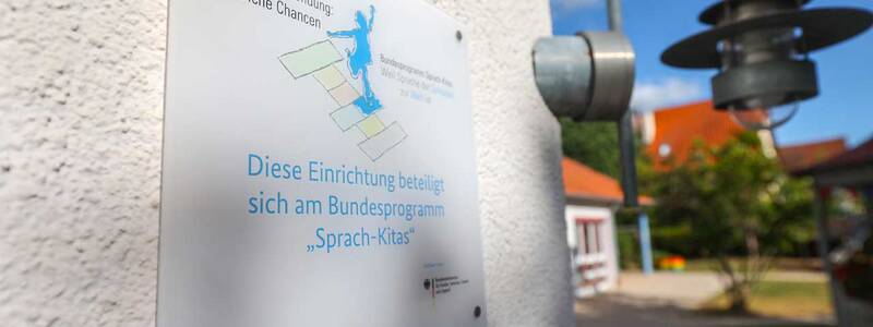 Eine weiße Tafel an einer Hauswand zeigt den Schriftzug „Diese Einrichtung beteiligt sich am Bundesprogramm Sprach-Kitas“. Daneben eine stilisierte Figur, die über bunte Felder hüpft. Im Hintergrund ist eine Lampe an der Wand und ein Gartenbereich mit Bäumen zu sehen.