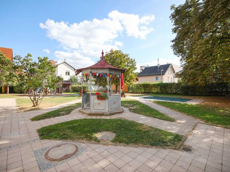 Ein kleiner Pavillon mit rotem Dach und Blumen, in der Mitte eines gepflasterten Gartens.
