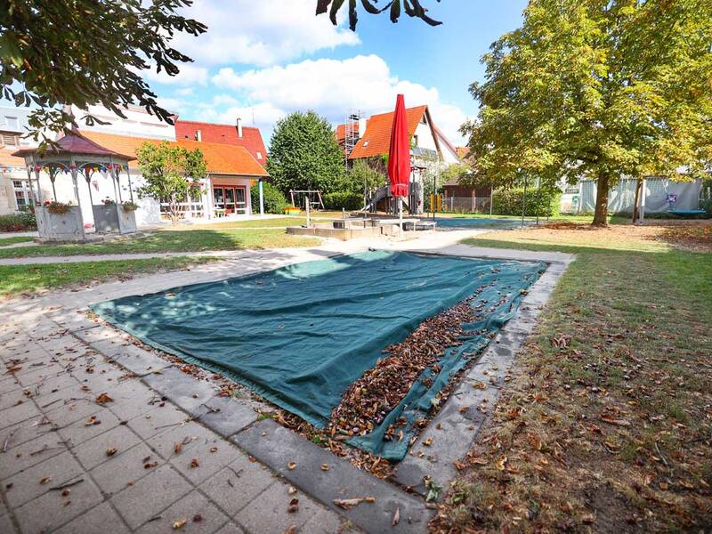 Im Vordergrund liegt ein großer, mit grünem Stoff abgedeckter Sandkasten. Darum herum sind gepflasterte Wege, Bäume mit herbstlichen Blättern und Spielgeräte wie Rutsche und Schaukeln zu sehen.