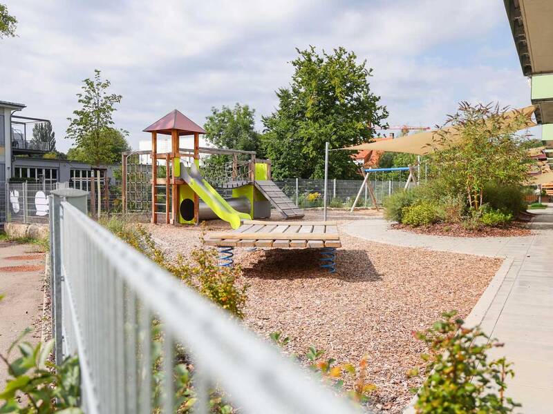Ein Spielplatz mit einem Klettergerüst, einer Rutsche und einer Wippe auf einem mit Holzschnitzeln bedeckten Boden neben einem modernen Gebäude.
