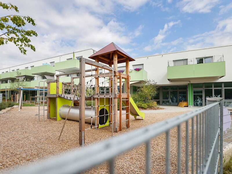 Ein moderner Spielplatz mit Klettergerüst und Rutsche befindet sich vor einem zweistöckigen Gebäude mit grünen Balkonen.